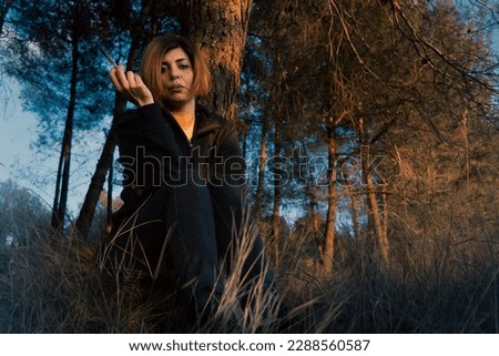 A woman in her 40s is smoking near a pine tree in a forest at sunset. She looks serious with a hint of contempt, as if tired and warning you to be careful