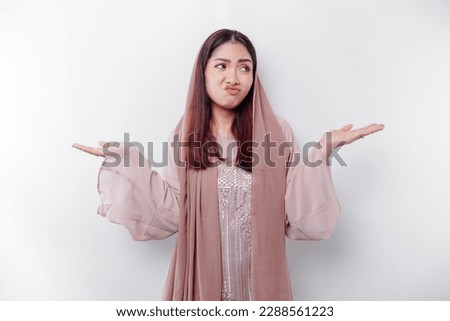 A thoughtful young Asian Muslim woman is wearing hijab and looks confused between choices, isolated by a white background