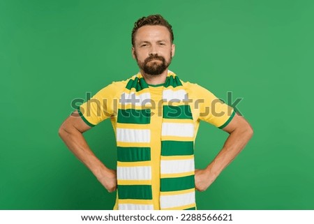 bearded soccer fan in striped scarf and yellow t-shirt standing with hands on hips isolated on green