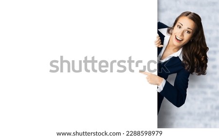 Happy excited woman in black confident suit showing pointing blank white banner signboard. Business and advertising concept. Copy space blank text area. White bricks loft wall background.