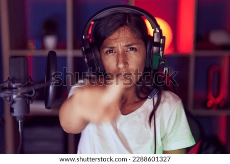 Young hispanic woman playing video games wearing headphones pointing with finger to the camera and to you, confident gesture looking serious 