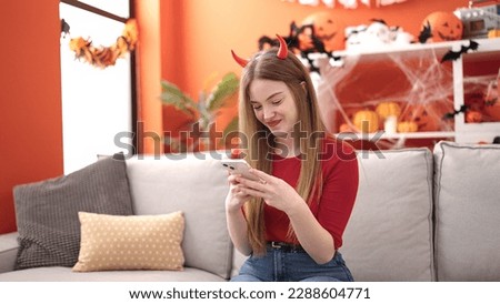 Young blonde woman smiling wearing devil costume using smartphone at home