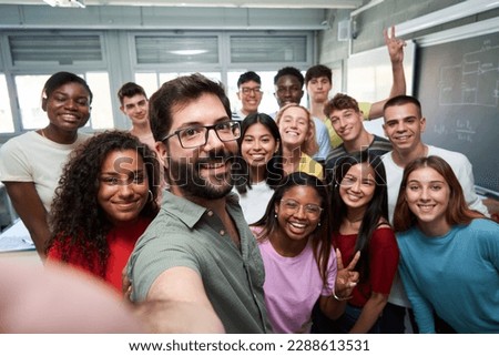 Happy selfie of young group of students taking a photo with their male teacher in the classroom, celebrating the end of course. Classmates from different countries, looking at camera with big smiles.