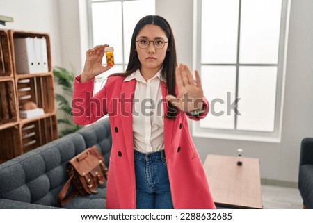 Young latin woman working as therapist holding jar of pills with open hand doing stop sign with serious and confident expression, defense gesture 