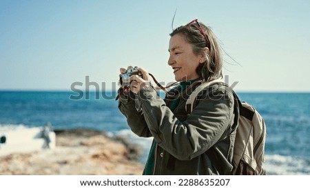 Mature hispanic woman with grey hair tourist wearing backpack holding vintage camera at seaside