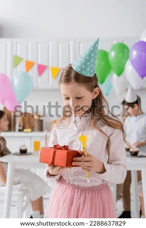 happy preteen girl holding birthday present and party horn near friends on blurred background Royalty-Free Stock Photo #2288637829