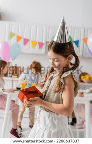 happy girl in party cap holding birthday present near friends on blurred background