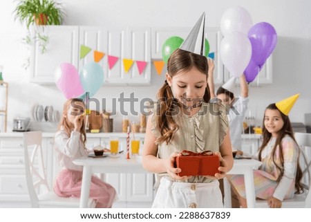 pleased girl in party cap holding birthday present near friends on blurred background