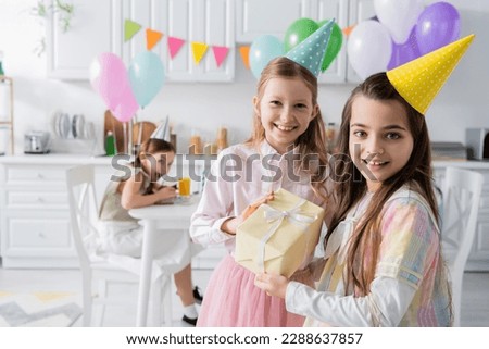 cheerful birthday girl in party cap receiving present from friend while looking together in camera Royalty-Free Stock Photo #2288637857