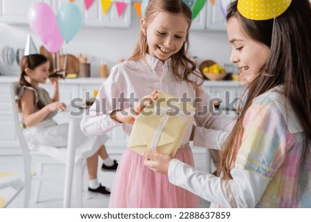 cheerful birthday girl in party cap receiving present from happy friend Royalty-Free Stock Photo #2288637859