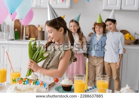 cheerful girl in party cap holding tulips next to friends during birthday party at home