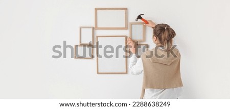 Young woman with hammer hanging blank photo frames on light wall