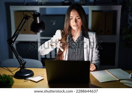 Young brunette woman working at the office at night with laptop looking unhappy and angry showing rejection and negative with thumbs down gesture. bad expression. 