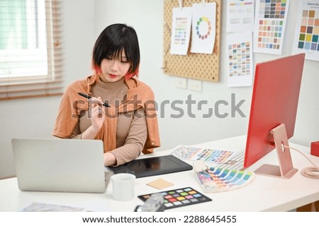 Charming young Asian female graphic designer using laptop, focusing on her new project, working in her studio.