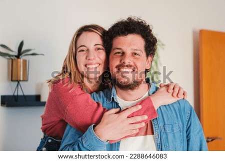 Close up portrait of heterosexual young caucasian couple embracing together at home living room, looking at camera with love attitude. Front view of married people hugging and smiling, relationship