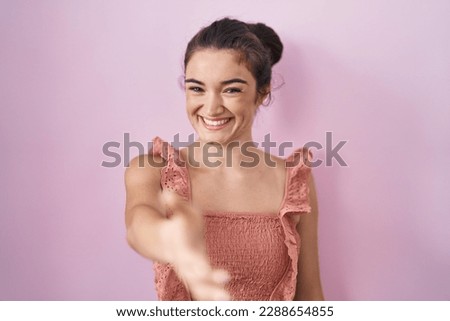 Young teenager girl standing over pink background smiling friendly offering handshake as greeting and welcoming. successful business. 