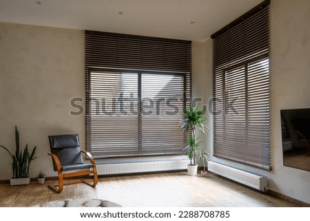 Wooden blinds on large windows in the interior. Living room with armchair and houseplants near windows with wood blinds. Motorized jalousie in the smart house. Royalty-Free Stock Photo #2288708785