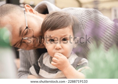 Dreamy picture of male infant staring looking at camera sitting on his father's legs with glasses in beer garden at the pub in Scotland, Edinburgh. Bokeh with plant. Multicultural family, mixed race.