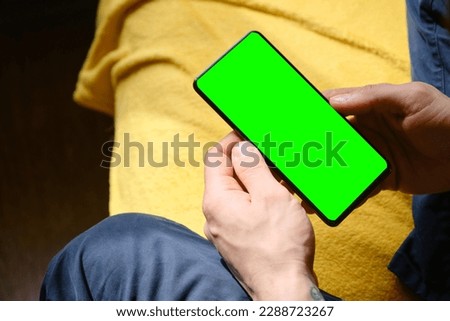 Guy Uses Green Mock-up Screen Smartphone. Young man sitting inside on yellow blanket on bed and uses phone to work or browse the Internet online, top view. Hands close-up.