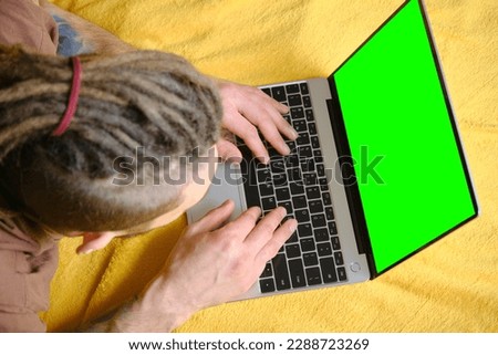 Guy Uses Green Screen notebook. Young man lying on yellow blanket on the bed and uses laptop to work or browse Internet online, typing text on keyboard, top view. Copy space for advertising or design.