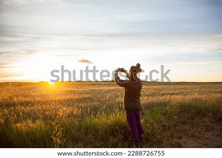 a boy from behind with long hair with a bow making photos with the mobile in the field looking at the sun in the sunset with the golden sky and with clouds