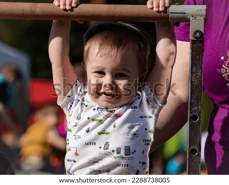 Young boy toddler smiling while hanging from gymnastics bar or playground equipment with mother beside