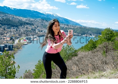 Young woman standing above a lake with stunning view and making selfie pictures with her mobile phone . 