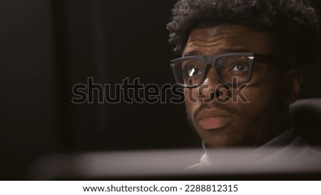 Face close up of African American man working or watching movie in studio with modern design. Film editor looks at monitors. Video footage reflects in the glasses. Freelance and remote work. Zoom in.