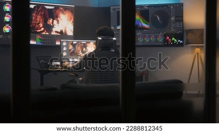 African American colorist does color grading in professional software on computer. Process of colour correction for video post production in modern studio. Multiple monitors with travel movie footage.