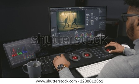 African American colorist makes color grading on control machine in modern studio. Big screens with action film footage and RGB colour correction graphic bar on the wall. Film post production process.