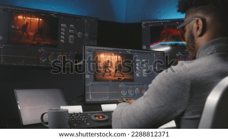 African American colorist makes color grading on control machine in modern studio. Big screens with action film footage and RGB colour correction graphic bar on the wall. Film post production process. Royalty-Free Stock Photo #2288812371