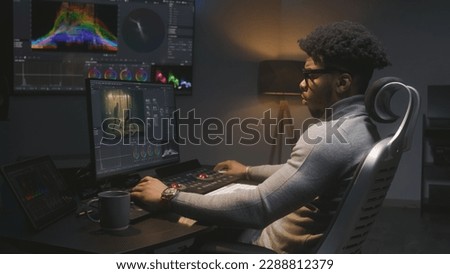African American film maker makes color grading on computer in modern studio. Big screen with RGB colour correction graphic bar on wall. Action film footage displayed on monitor. Colorist equipment.