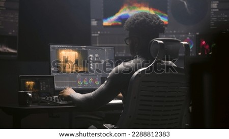 African American film editor does color grading on computer in modern studio. Big monitors with RGB colour correction graphic bar and program interface on the wall. Video post production. Handheld. Royalty-Free Stock Photo #2288812383