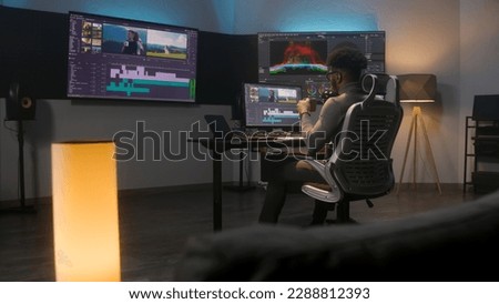 African American multimedia artist drinks coffee and makes color grading in editing program on computer. Process of video post production in modern studio. Multiple monitors with travel movie footage.