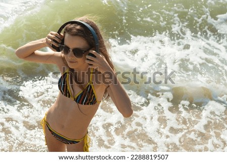 Pretty young girl in bikini with black headphones in her hands on beautiful wavy sea background.