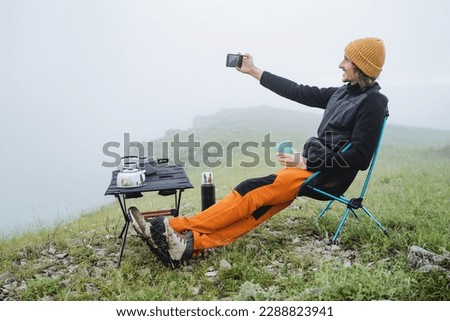 Take a selfie at lunch, a guy takes pictures of himself in nature sitting in a chair, online communication outside the house, going to a social network in nature, a man in the mountains is buzzing