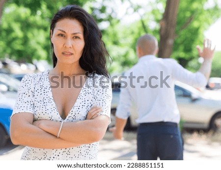 Woman and man having quarrel. Woman standing with folded arms on her chest and man brushing off in background.