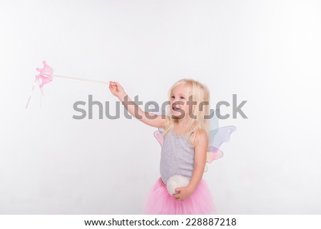 Half-length portrait of little fair-haired lovely smiling girl wearing pretty grey vest pink skirt and white wings standing aside conjuring over someone. Isolated on white background