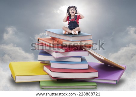Asian boy listening to the music and sitting on pile of books 