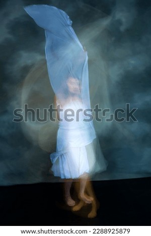 Mystical, blurred image of an adult female professional dancer in a white dress, dancing with a floating, blue, gauzy fabric in a studio, shot against a gray background.
