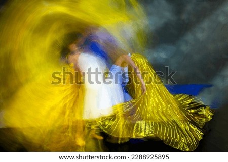 Mystical, blurred image of a female professional dancer in a white dress, dancing with a floating, shiny gold fabric in a studio shot, against a gray background.