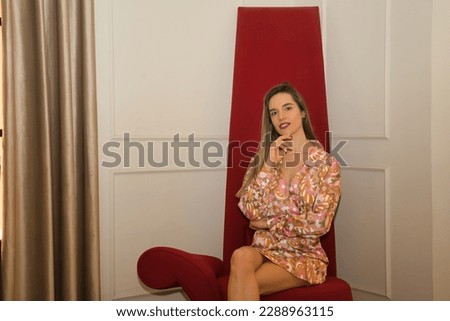 Young, beautiful, blonde woman, with flower dress, sitting on a red designer armchair, independent and empowered. Concept of empowerment, independence, security.