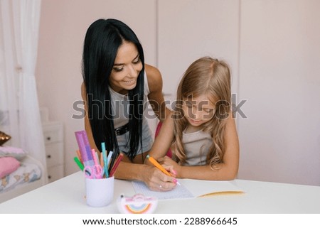 Mom helps her daughter do her homework at home. They look carefully at the notebook where the girl is writing.