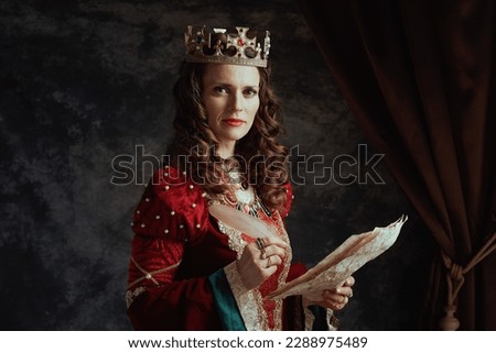 medieval queen in red dress with parchment and crown on dark gray background. Royalty-Free Stock Photo #2288975489