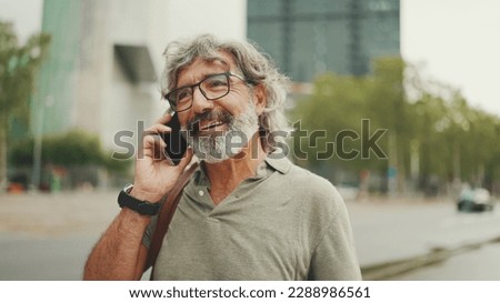 Friendly middle aged man with gray hair and beard wearing casual clothes using his mobile phone. Mature gentleman in eyeglasses talking on cell phone outdoors Royalty-Free Stock Photo #2288986561