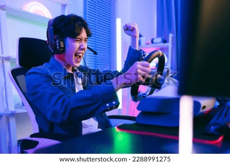 Asian male pro gamer playing an online car drive simulation game with steering wheel controller. Gamer winning an sports game with victory emotion. The player rejoices in victory in the competition.