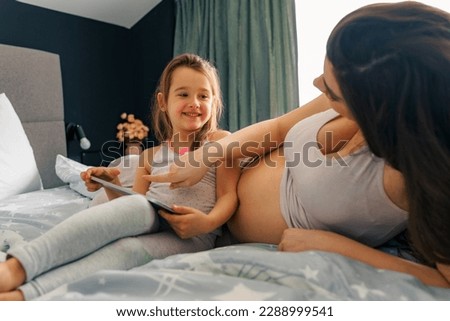 Mom and daughter sitting on a bed and bounding before new baby arrives, watching cartoons on tablet placed on daughter's lap