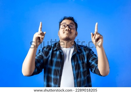One Indian young man looking up and pointing with both fingers, presenting copy space, isolated on blue background