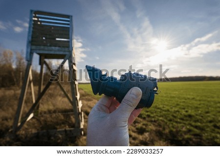 Thermal night vision device in hand against the background of a hunting tower on a field on a sunny day.