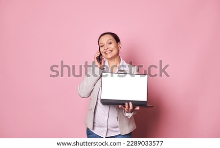 Pleasant pregnant woman, sales manager, entrepreneur talking on mobile phone and showing at camera a laptop with white screen with copy space for mobile apps, smiling isolated over pink background
